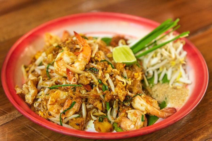 A plate of shrimp pad thai garnished with fresh bean sprouts, a lime wedge, and garlic chives, served on a red plate on a wooden table.