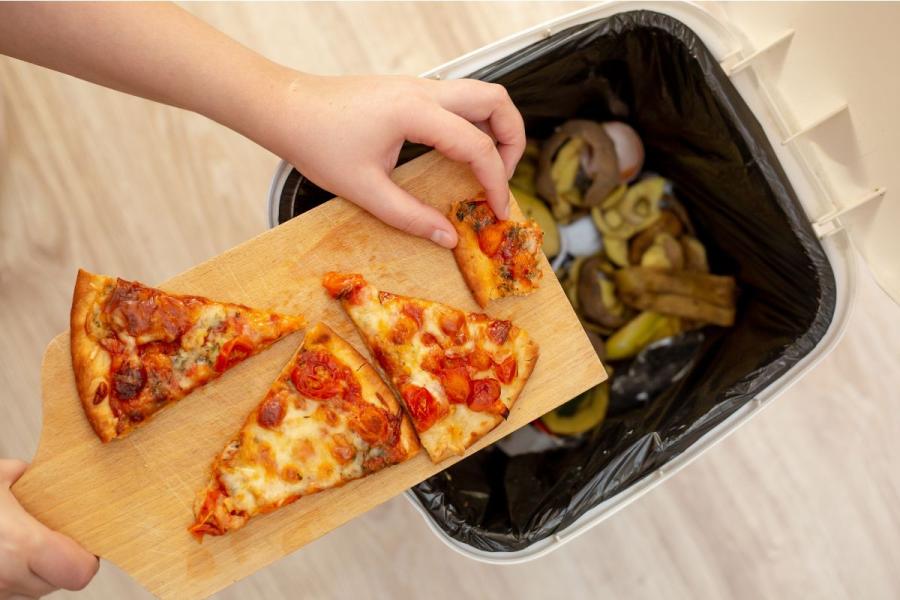A hand scraping three uneaten slices of tomato pizza off a wooden board into a kitchen bin already filled with discarded vegetable peels and food scraps.