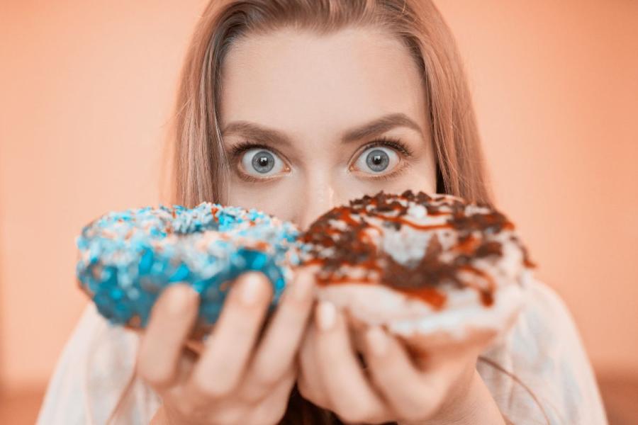 A wide-eyed young woman holding two frosted doughnuts up in front of her face — one topped with blue sprinkles, the other with chocolate drizzle.