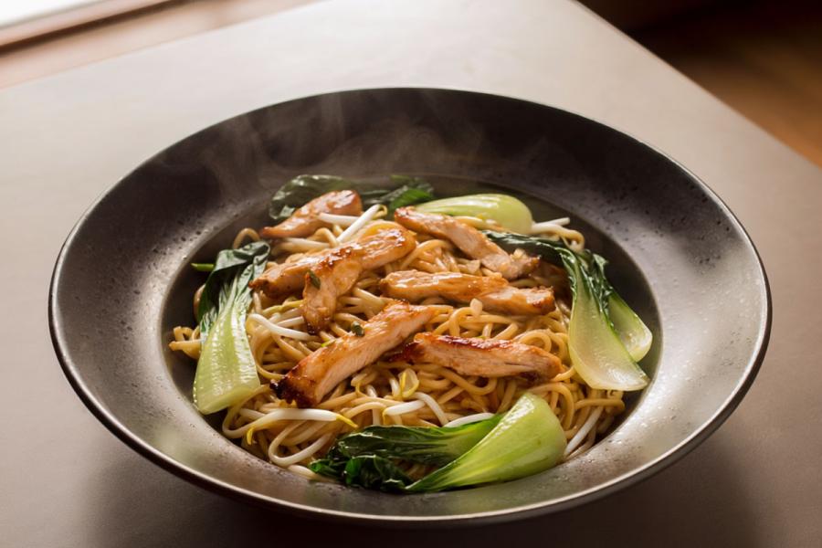 Steaming bowl of wok-fried noodles topped with sliced glazed chicken, bok choi, and bean sprouts. This is a synthetic image.