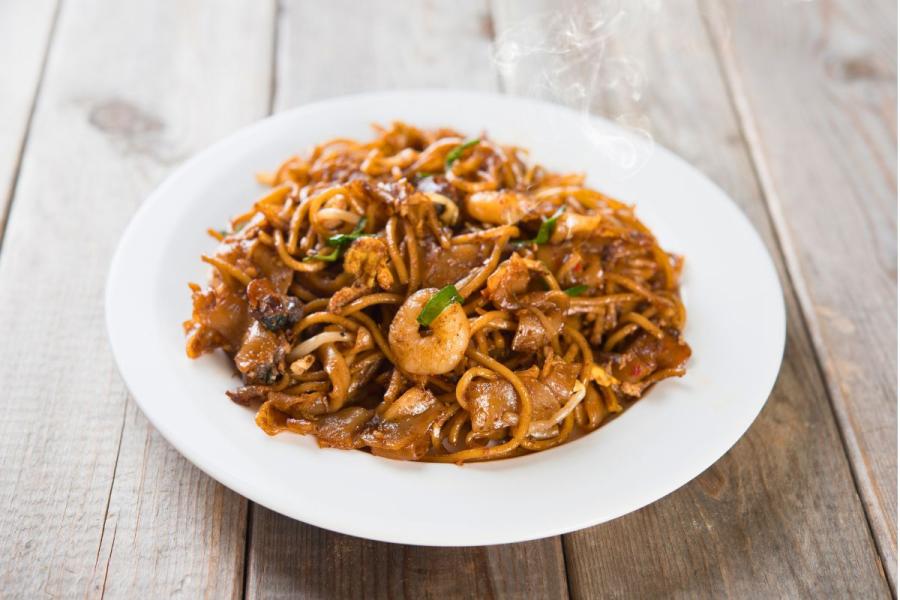 A plate of char kway teow — stir-fried flat rice noodles with shrimp, egg, bean sprouts and scallions, glistening with dark soy sauce and lightly charred, served on a white plate on a wooden table, with steam rising.