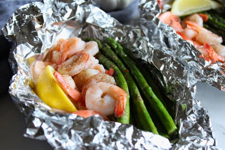 A foil packet filled with cooked shrimp, asparagus spears and lemon wedges, seasoned with black pepper.