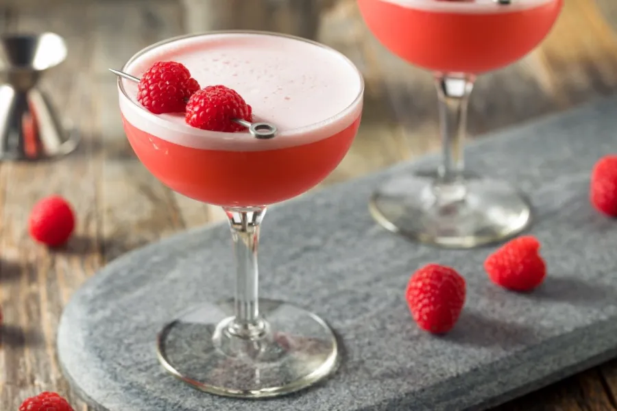 A pink, foam-topped cocktail in a coupe glass garnished with two raspberries on a pick, on a gray stone board with fresh raspberries scattered around.