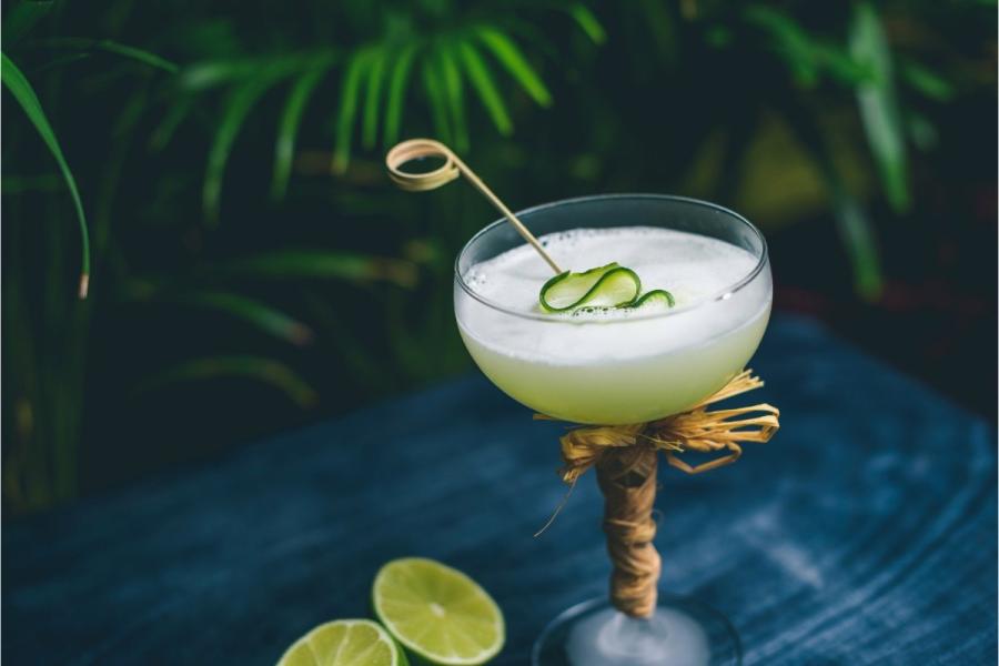 A pale green, frothy cocktail in a raffia-wrapped coupe glass, garnished with a curl of cucumber on a pick, on a blue wooden surface with lime halves and tropical foliage behind.