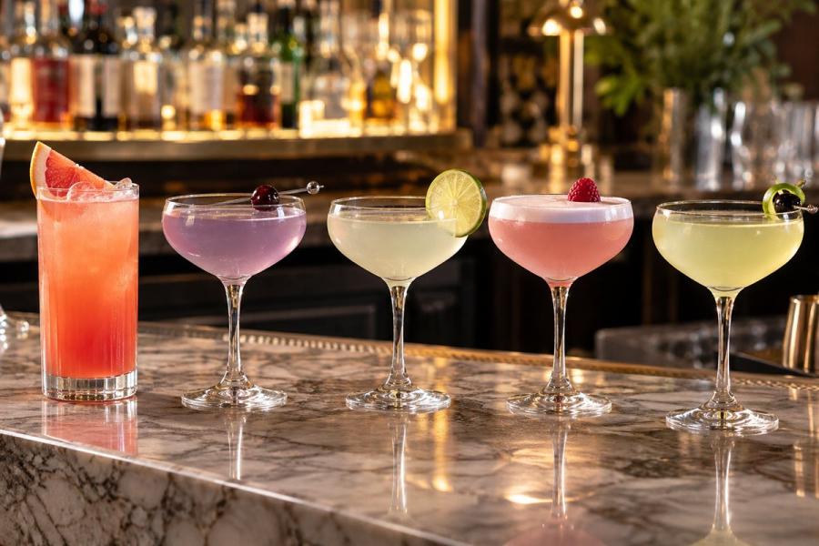 Five colorful cocktails in a row on a marble bar top — a tall grapefruit highball, a lavender coupe with a cherry, a pale green coupe with a lime wheel, a pink foamy coupe with a raspberry, and a yellow coupe with an olive — against a warmly lit back bar.