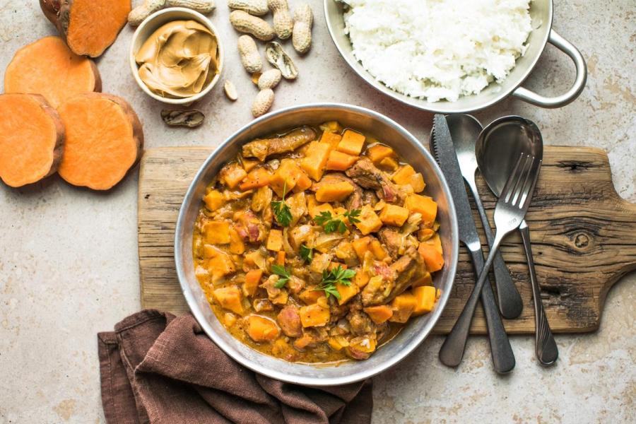 A bowl of chicken and sweet potato peanut stew in a rich orange sauce, garnished with parsley, on a wooden board surrounded by sweet potato slices, peanuts, a dish of peanut butter, and a bowl of white rice.