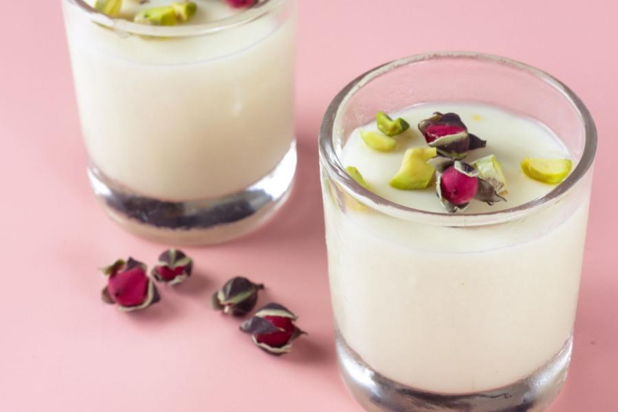 Two glasses of mahalabiya, a pale Egyptian milk pudding, topped with chopped pistachios and dried rose buds, on a pink background