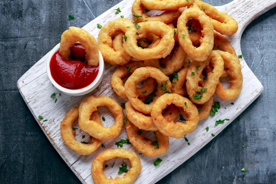 A pile of golden battered onion rings on a white wooden board, sprinkled with parsley, with a small bowl of ketchup