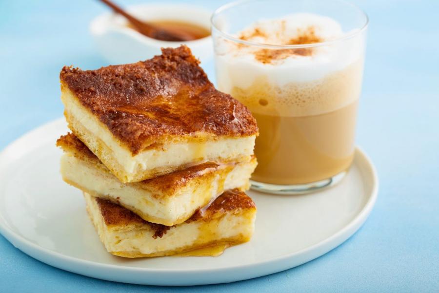Stack of sopapilla cheesecake bars drizzled with honey, accompanied by a frothy cinnamon-topped coffee.